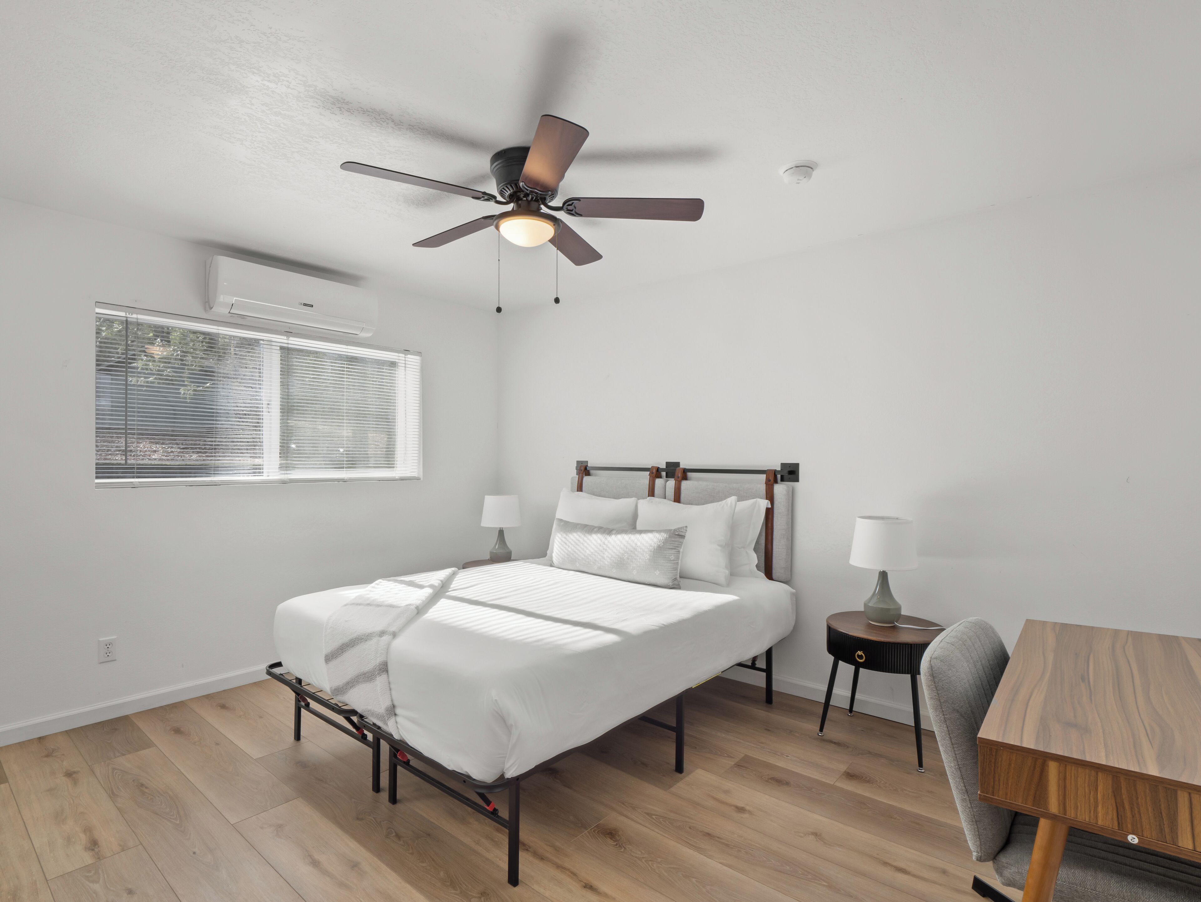 Un dormitorio con cama, mesitas de noche, un escritorio, un ventilador de techo y una ventana con persianas.