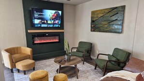 Een woonkamer met een flatscreen televisie, een open haard, groen en bruin meubilair en een decoratief wandobject.