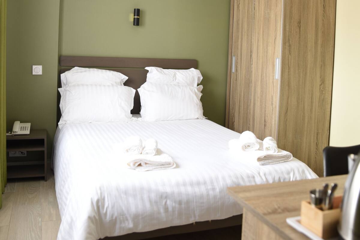 A neatly made bed with white linens, a wooden wardrobe, and a bedside table with a phone and a kettle.