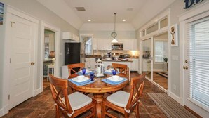A spacious kitchen with white cabinets, a dining area with wooden chairs and blue placemats, and a large window with blinds. - Seaglass Village 805: Coastal Charm Cottage - Spacious Port Model w/ Resort Amenities (Wells)