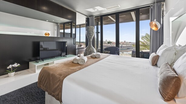 A modern hotel room with a large bed, a flat-screen TV, a vase, and a view of the city. - Penthouse Grand Noir | Outdoor Jacuzzi | Near the Strip (Albufeira)