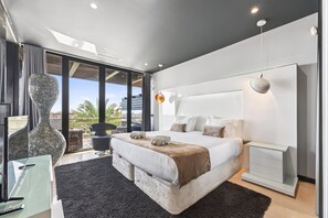 Ein modernes Schlafzimmer mit einem großen Bett, einem Glastisch und Blick auf Palmen.