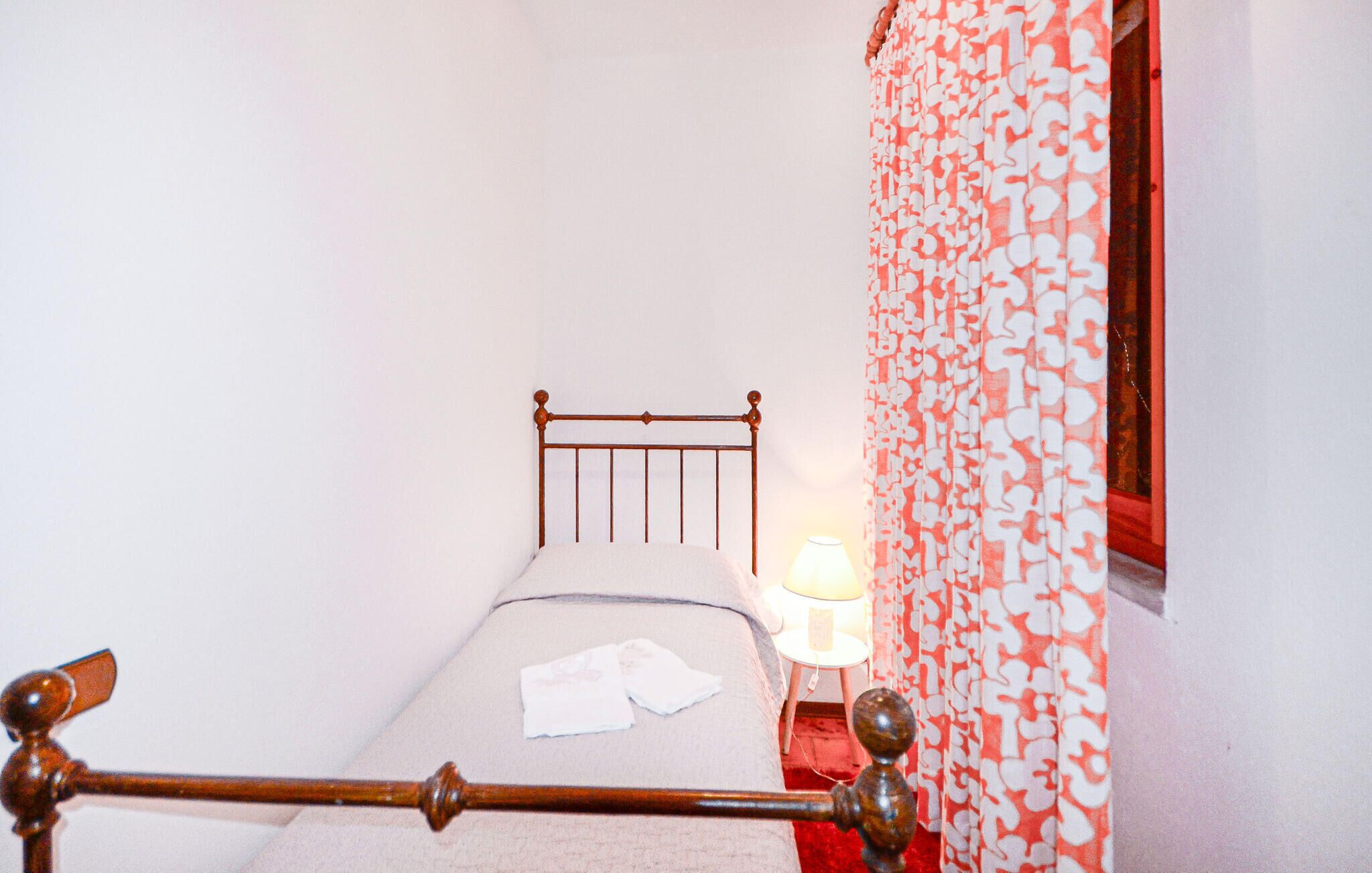 A small, narrow room with a single bed, a nightstand with a lamp, and a patterned curtain.