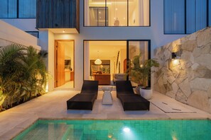 A modern outdoor pool area with lounge chairs, a stone wall, and a view into the interior of the building. - Villa Santai Santai by Alfred in Bali (Canggu)