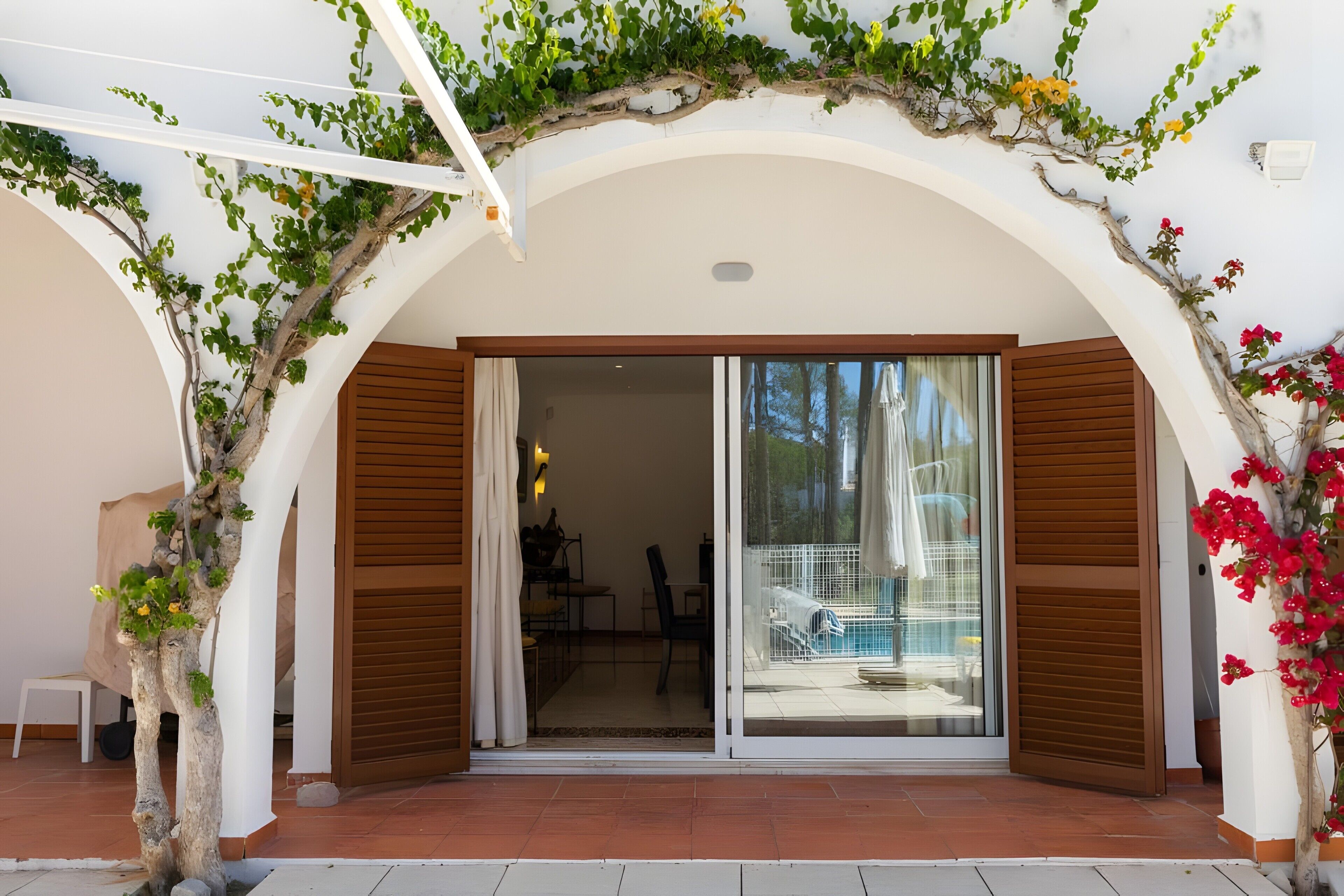 A covered outdoor area with a sliding glass door leading to a pool, surrounded by white walls and wooden shutters.