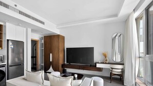 Un salon moderne comprenant un canapé, un téléviseur à écran plat, un coin repas avec une table en bois et une cuisine équipée d’un réfrigérateur.