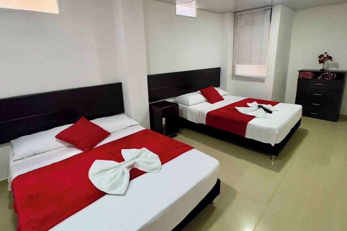 A hotel room with two beds, each with red and white bedding, a small nightstand, a dresser, and a window with blinds.