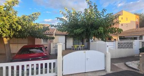 A residential area with a red car parked in front of a white gate, a yellow building, and a tree providing shade. - Lovely semi-detached villa in residence with pool near Richelieu beach, Cap d'Agde (LE CAP D AGDE)