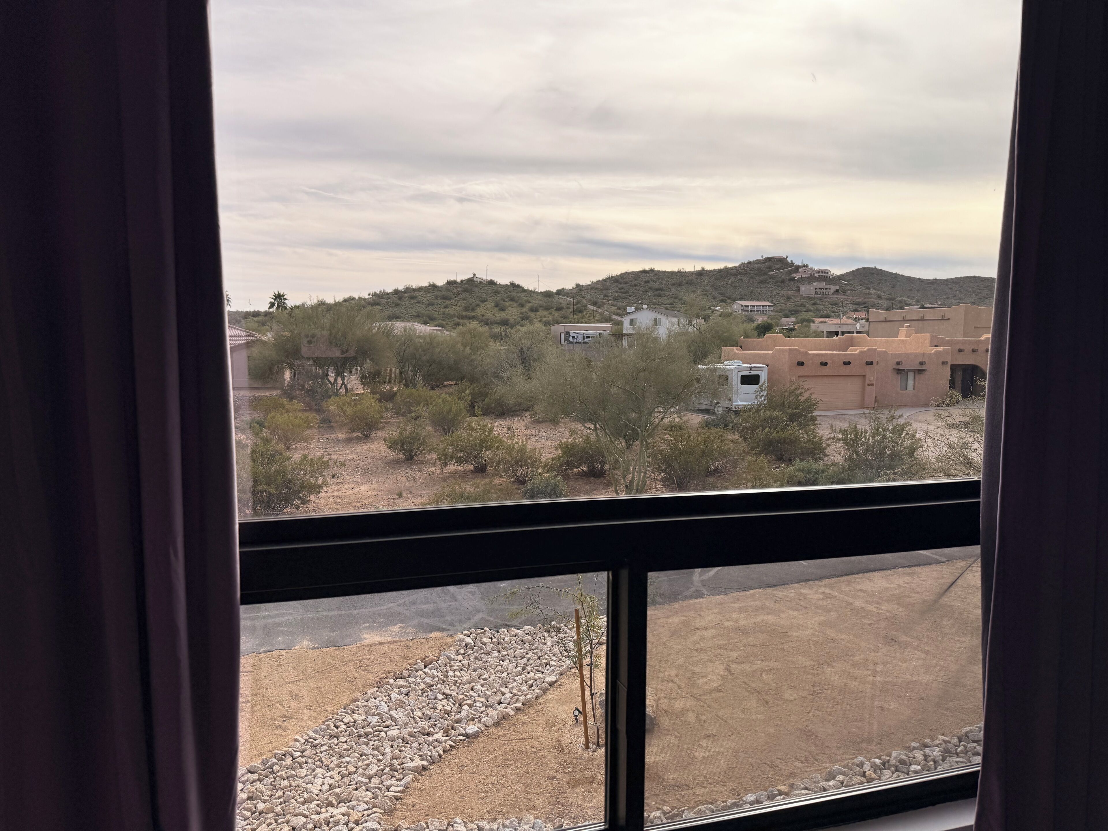 A view from a window with curtains, featuring a desert landscape with sparse vegetation, a few buildings, and a distant hill.