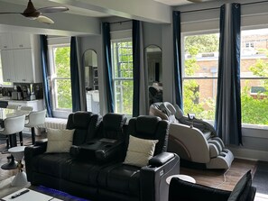 A modern living room with a sofa and massage chairs, large windows with curtains, a dining area, and a kitchen. - 8BR • 2 Kitchens • Walk to UT • Huge TV • Massage (Austin)
