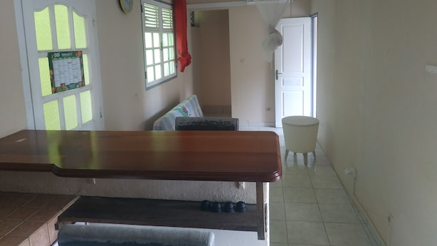 A room with a wooden bench, a tiled floor, a bed with a floral bedspread, a wall-mounted clock, and a calendar. - Appart close to everything. (Gros-Morne)