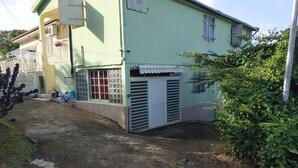 A two-story building with a green facade, a covered porch, and a gated entrance. - Appart close to everything. (Gros-Morne)
