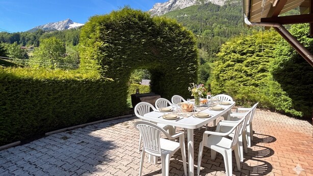 Zona pranzo all'aperto con tavolo apparecchiato per quattro, circondata da sedie bianche e vista su un paesaggio montuoso.