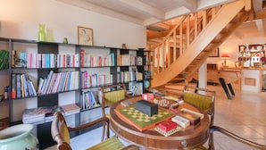 Un'ampia stanza con una scala in legno, un tavolo rotondo con scacchi e scaffali colmi di libri.