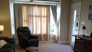 Ein Wohnzimmer mit einer schwarzen Ledersofa, einem schwarzen Ledersessel, zwei runden Holztischen, einem beigen Teppich und einem großen Fenster mit Vorhängen.