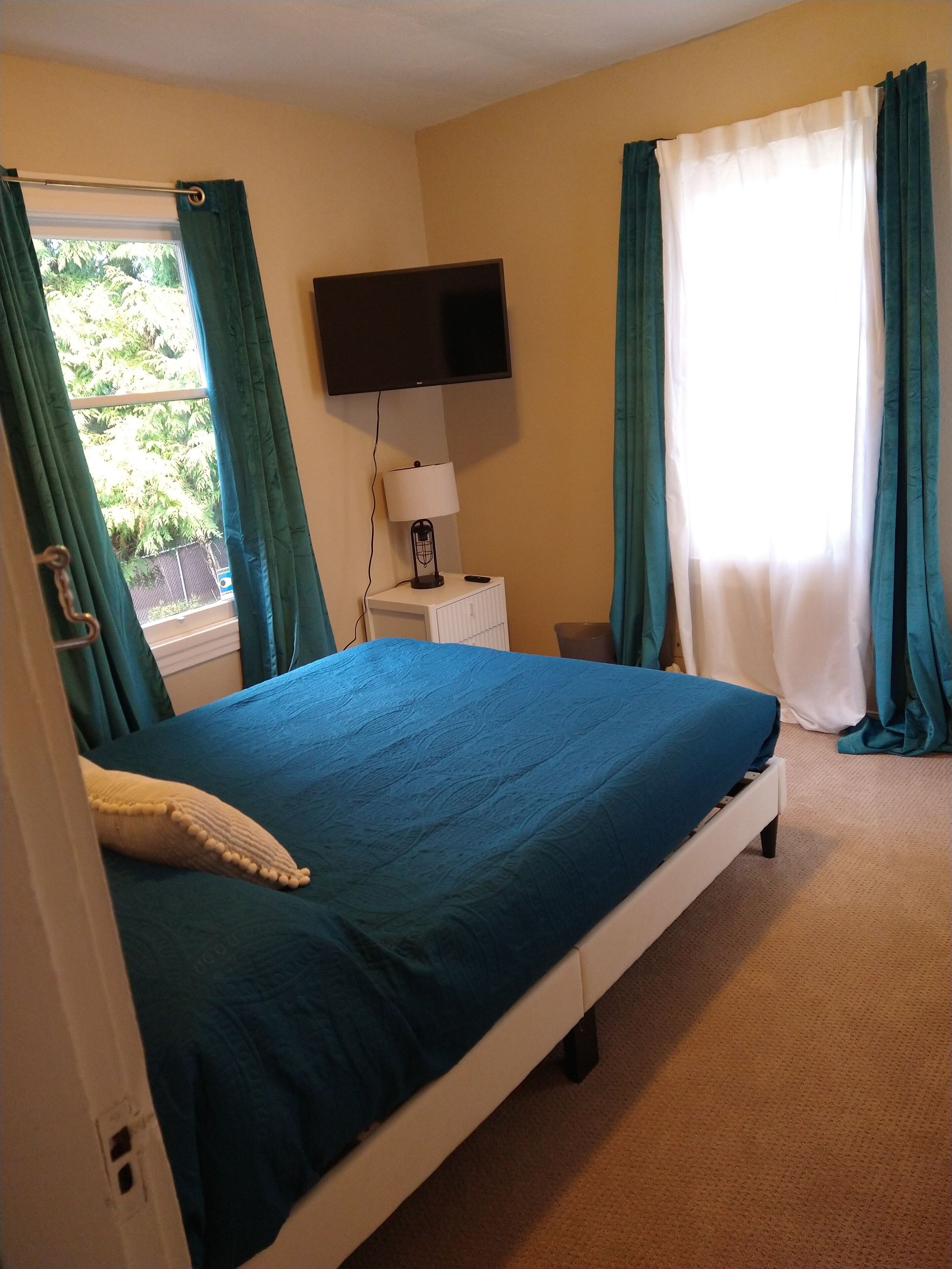 Une chambre à coucher avec un lit, un téléviseur fixé au mur, une table de chevet avec une lampe et des rideaux aux fenêtres.
