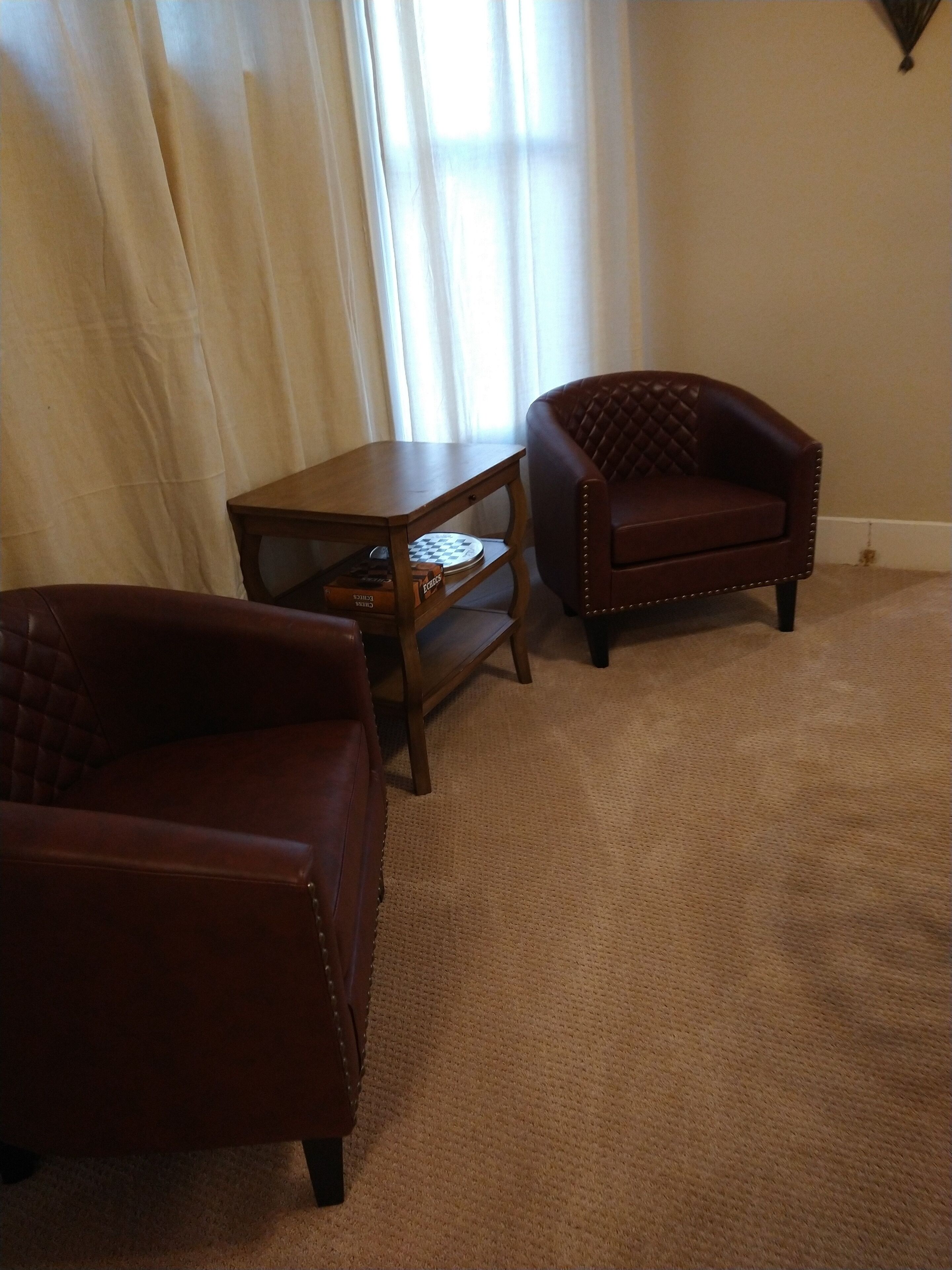 Una habitació amb una butaca de cuir marró, una taula auxiliar de fusta amb un tauler d'escacs i una finestra gran amb cortines de tela fina.