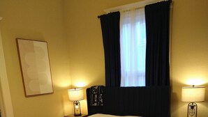 Ein ordentlich bezogenes Bett mit weißer Bettdecke, zwei Nachttischlampen, einem dunklen Kopfteil, einem Fenster mit dunklen Vorhängen und einem gerahmten Bild an der Wand.