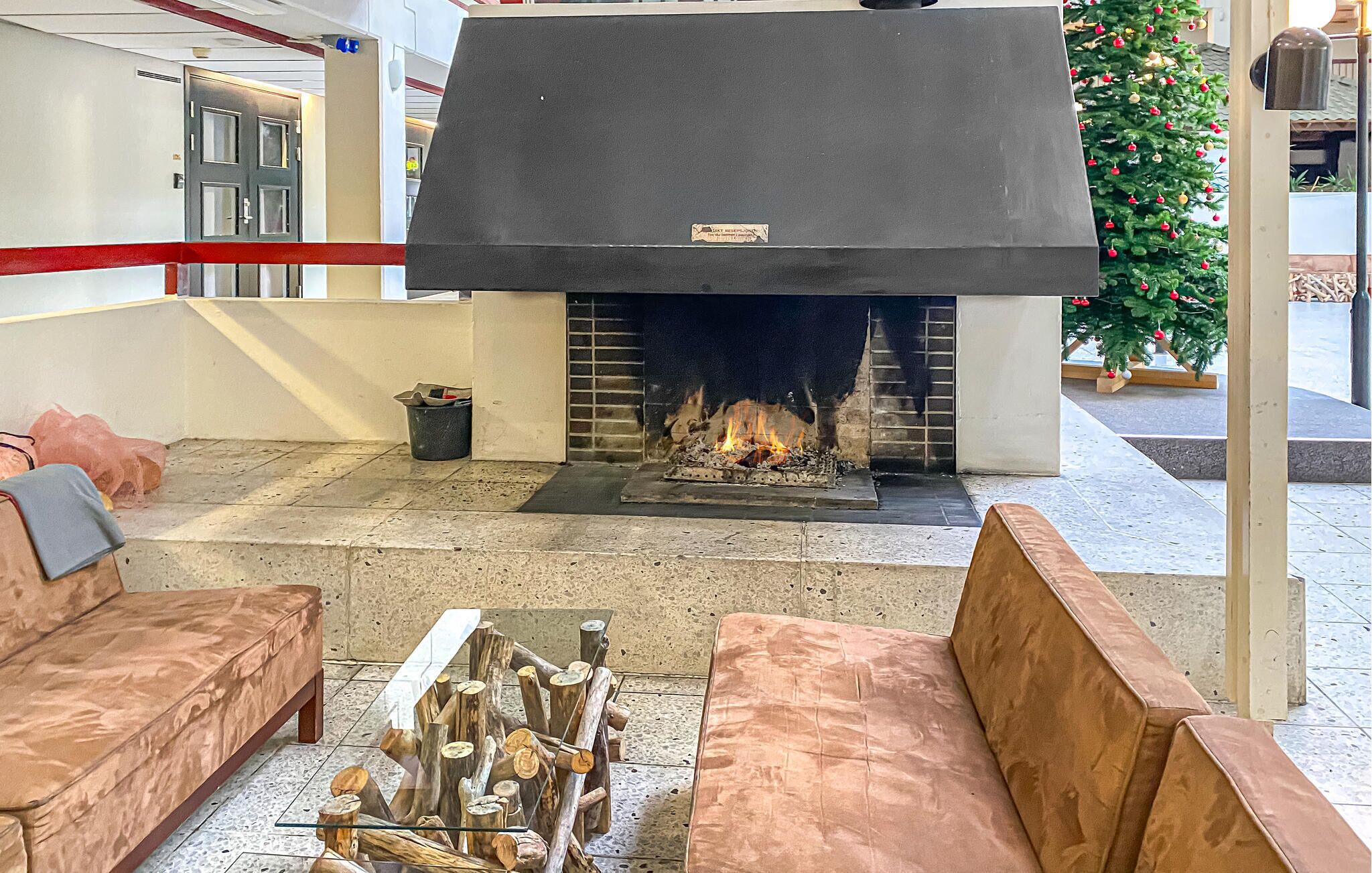 Une cheminée avec des bûches, un canapé et un sapin de Noël dans un espace de réception.