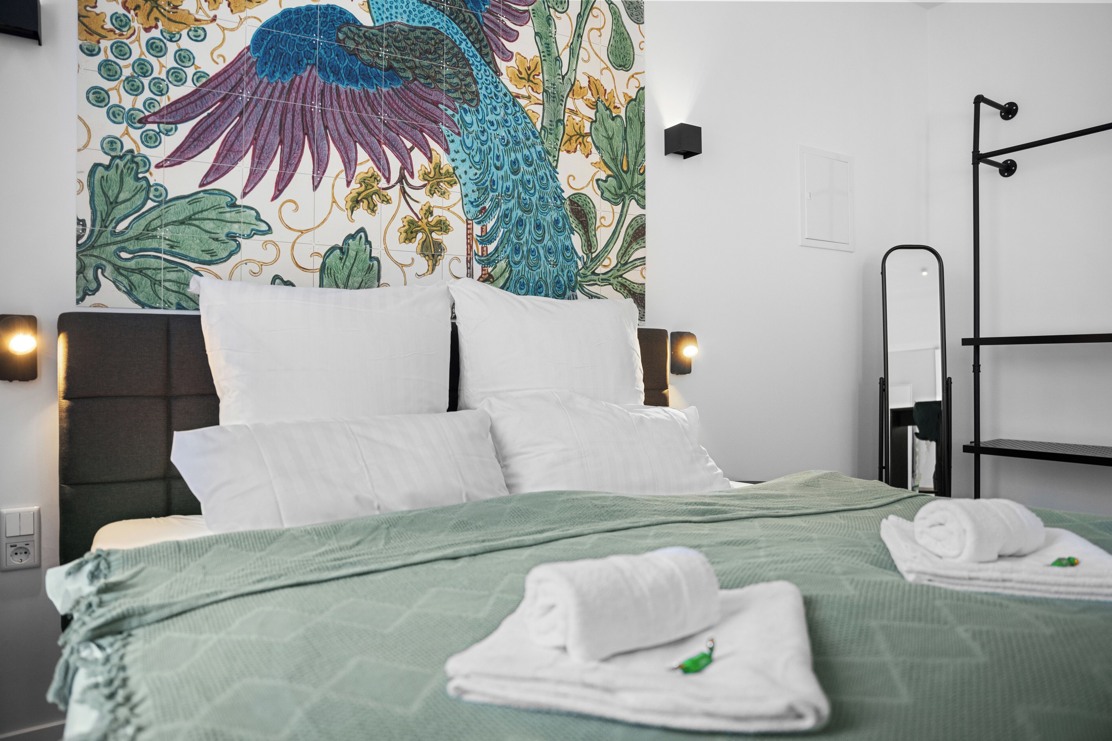 A neatly made bed with white linens and a green bedspread, a wall tapestry with a bird and floral design, and a black metal rack with towels.