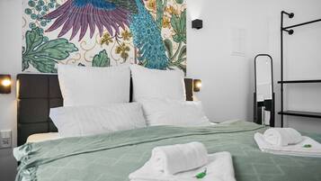 A neatly made bed with white linens and a green bedspread, a wall tapestry with a bird and floral design, and a black metal rack with towels.