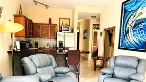 A living room with two blue reclining chairs, a wooden coffee table, and a kitchen area with a toaster and refrigerator.