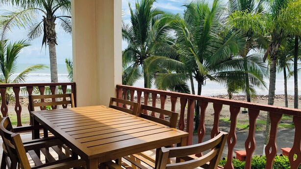 A wooden table and chairs on a balcony with a view of palm trees and the beach.
