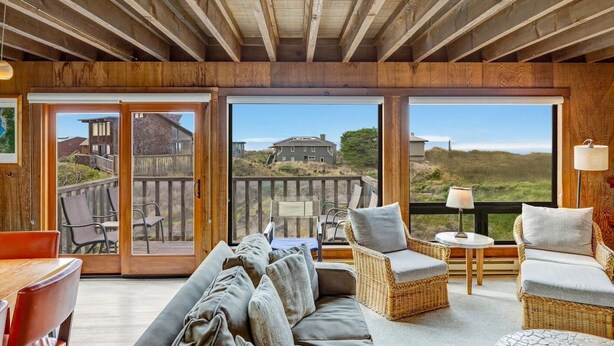 A cozy room with a wooden ceiling, a large sliding glass door leading to an outdoor deck, wicker furniture, and a view of the landscape. - House 25 | 4 Bedroom (Watsonville)