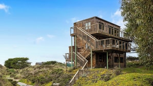 A multi-level wooden building with a staircase leading to the upper floors, surrounded by natural vegetation and a clear sky. - House 25 | 4 Bedroom (Watsonville)