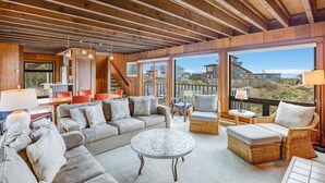 A spacious living area with a large sofa, wicker chairs, and a round coffee table. A staircase leads to an upper level with a balcony overlooking the landscape. - House 25 | 4 Bedroom (Watsonville)