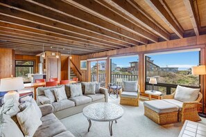 A spacious living area with a large sofa, wicker chairs, and a round coffee table. A staircase leads to an upper level with a balcony overlooking the landscape. - House 25 | 4 Bedroom (Watsonville)