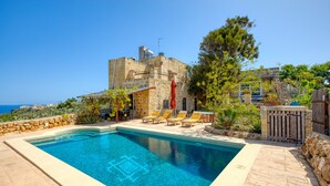 Una piscina rectangular con agua azul cristalina, rodeada de muros de piedra y un edificio de piedra con balcón.