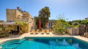 Una piscina con sillas de descanso y una pérgola, rodeada de edificios de piedra y follaje.