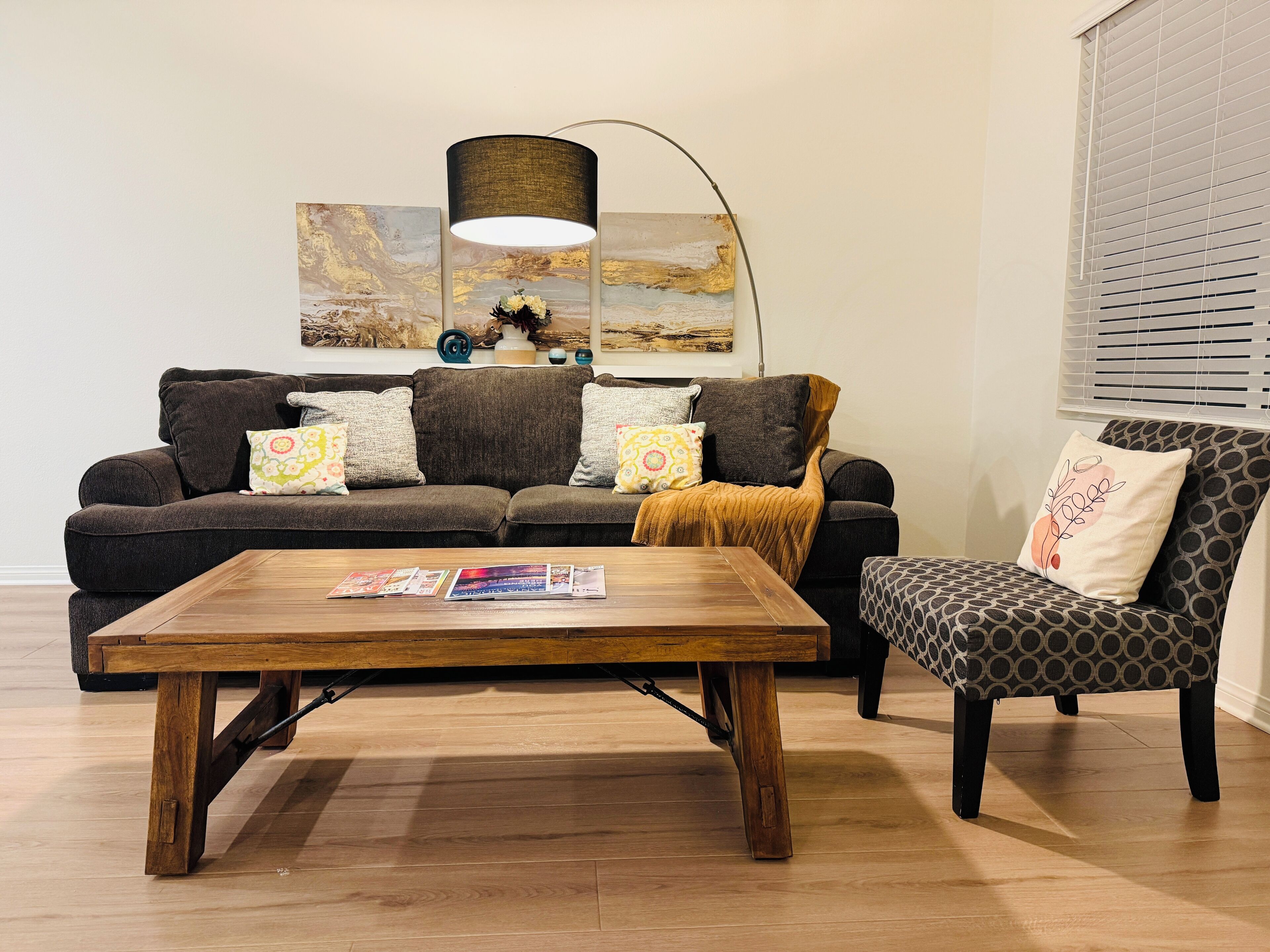 Ein Wohnzimmer mit Couch, Sessel und Holztisch. Bunte Kissen und eine Stehlampe sorgen für Gemütlichkeit.