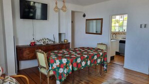 A dining area with a table covered in a floral tablecloth, wooden chairs, a wooden cabinet, a television mounted on the wall, and a window with a view of greenery outside. - Reñaca Village, in Viña del Mar, 3 min walking to beach, @casavista_panoramica (Viña del Mar)
