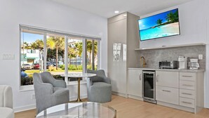 A modern kitchen with a built-in oven, a coffee maker, and a TV mounted on the wall displaying a beach scene. - Beachside Bliss • Renovated Studio • Walk to Ocean & Restaurants + Saltwater Pool (Fort Lauderdale)