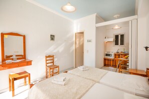 Ein Schlafzimmer mit zwei Betten, einem Holzstuhl, einem kleinen Tisch, einem Spiegel und einer Küchenzeile im angrenzenden Raum.