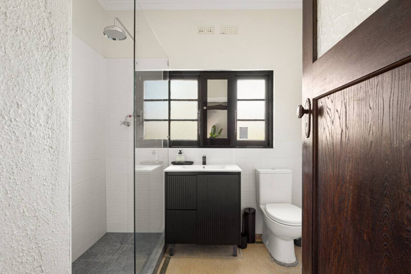 Een badkamer met een douchecabine van glas, een wit toilet, een donkere wastafel en een raam boven de wasbak.