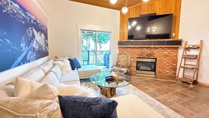A modern living room with a white sofa, a glass coffee table, a fireplace, and a large TV mounted on the wall. - Summit View II | Ski-in · Hot Tub · Sleeps 6 (Kimberley)