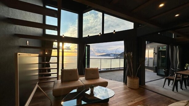 A spacious living area with a large window overlooking a body of water, a modern sofa set, and a dining area. - VILLA IZU Zessei (Ito)