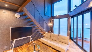 A modern living room with a large window, a sofa, a TV on a stand, and a staircase. - VILLA IZU Zessei (Ito)