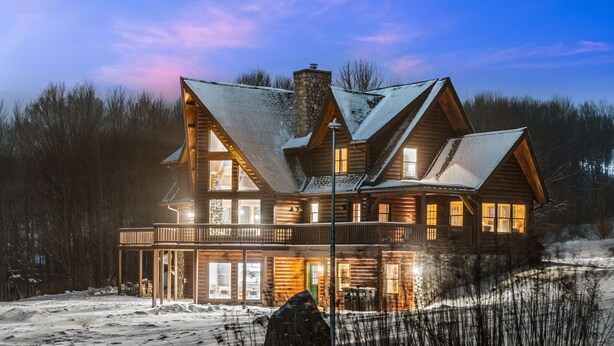A large, multi-story log cabin with a prominent chimney, multiple windows, and a balcony.