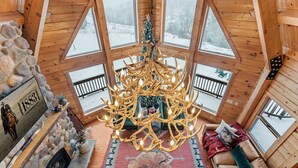 A cozy cabin interior with a stone fireplace, a chandelier, and a view of a snowy landscape through large windows.