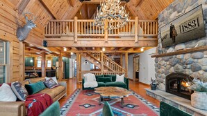 A spacious log cabin interior with a high ceiling, a stone fireplace, and a staircase leading to an upper level.
