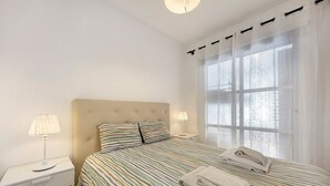 Un dormitorio con cama, dos mesitas de noche, una ventana con cortinas y cabecero.