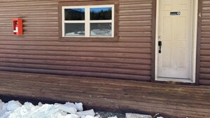 A wooden cabin with a window and a door, a fire extinguisher mounted on the wall, and a snow-covered ground.