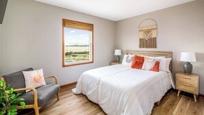 Ein Schlafzimmer mit einem großen Bett, zwei Nachttischen, einem Sessel, einem Fenster mit Blick auf ein Feld und einem Wandbild.