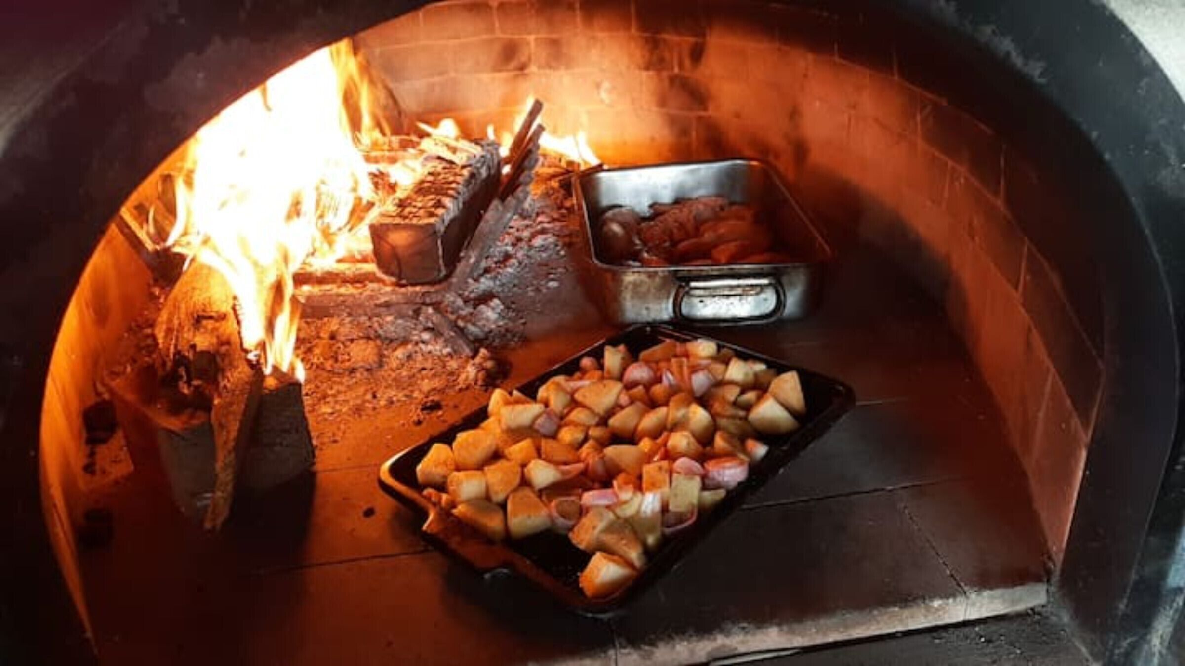 A pizza oven with wood burning, a tray of sliced potatoes, and a metal container with meat.