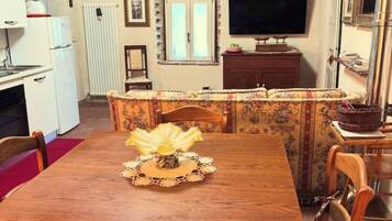 A dining area with a wooden table, chairs, a television, and a kitchen area with a refrigerator and stove.