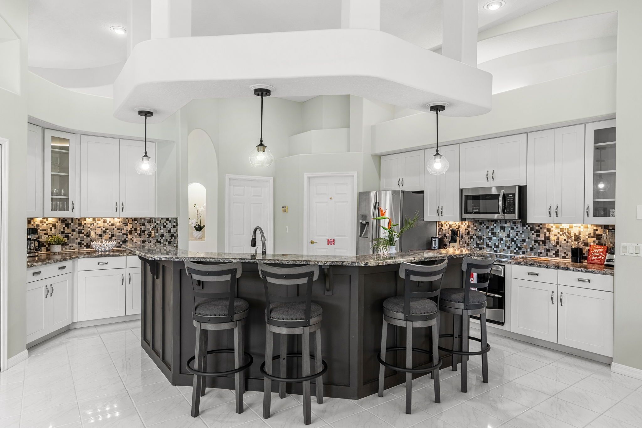 A modern kitchen with a central island, bar stools, white cabinets, and a tiled backsplash.